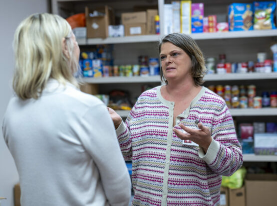 Two women discussing assistance program in front of shelves of donated food